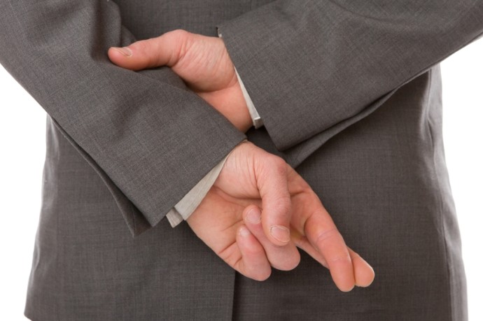 Close-up photo of a man, dressed in a business suit, crossing his fingers behind his back, implying fraud.
