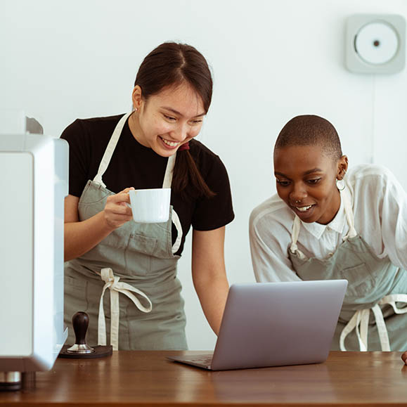 Two smiling women in aprons, lookin at an open laptop.