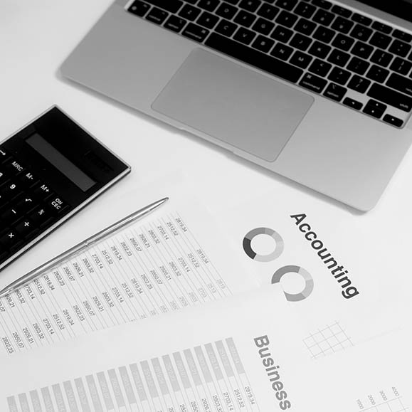 Black and white photo of papers that say "Accounting" and "Business" on them, with data and a couple circle ring graphs depicting data, in front of a laptop.