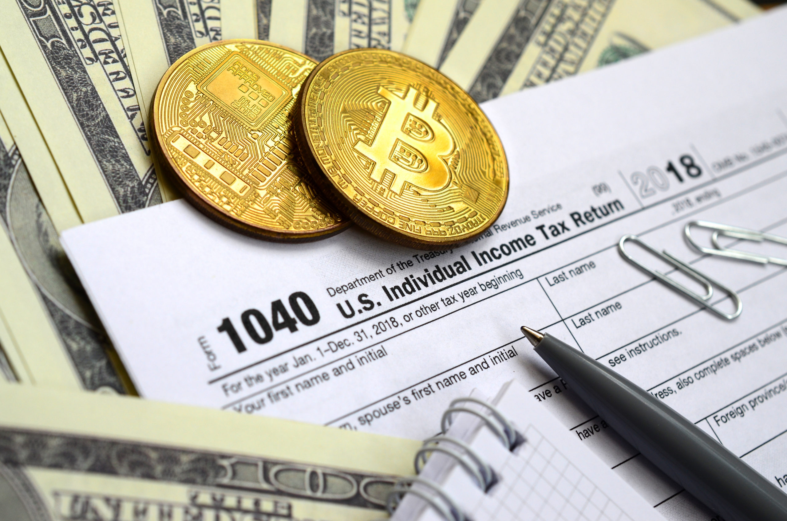 Photo of a pen, physical bitcoins and $100 bills on a 1040 U.S. tax form.